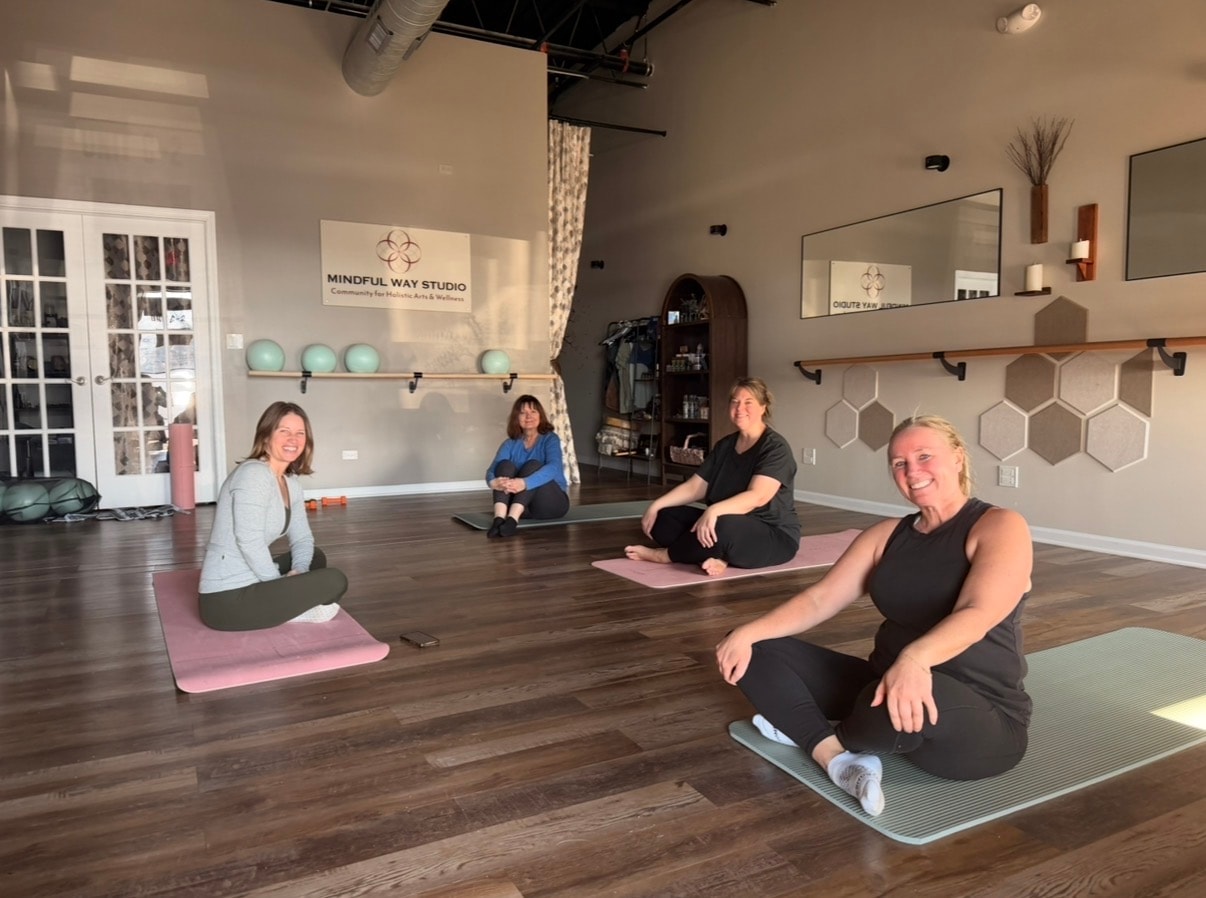 Group fitness class at Mindful Way Studio Mokena Illinois — four women smiling on yoga mats with Mindful Way Studio sign visible